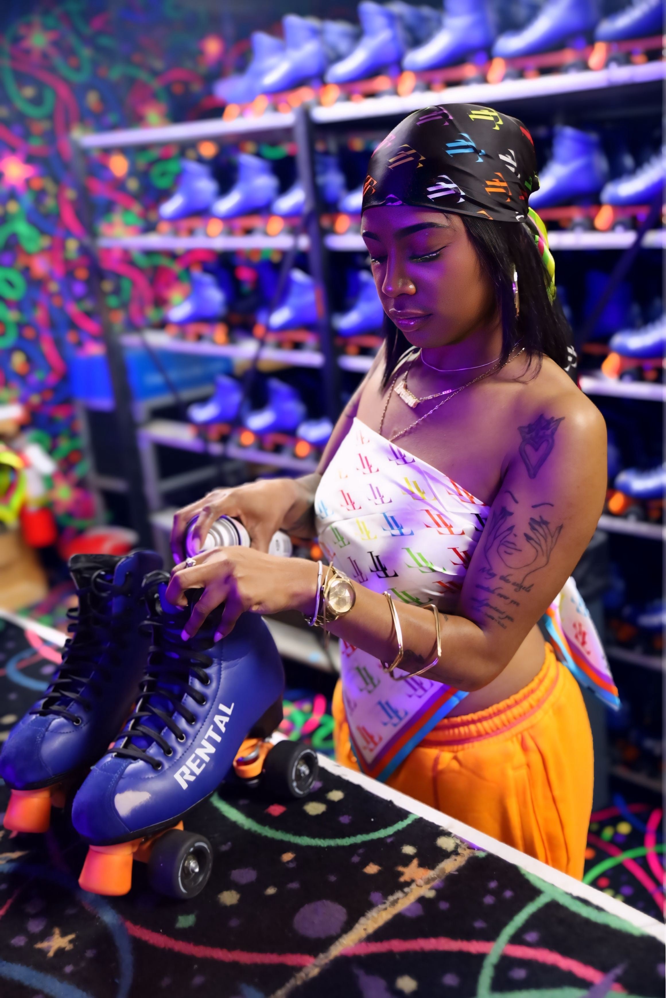 A woman in a roller skate shop applies polish to a blue Rival roller skate while wearing a colorful patterned top, orange sweatpants, and a black silk scarf.